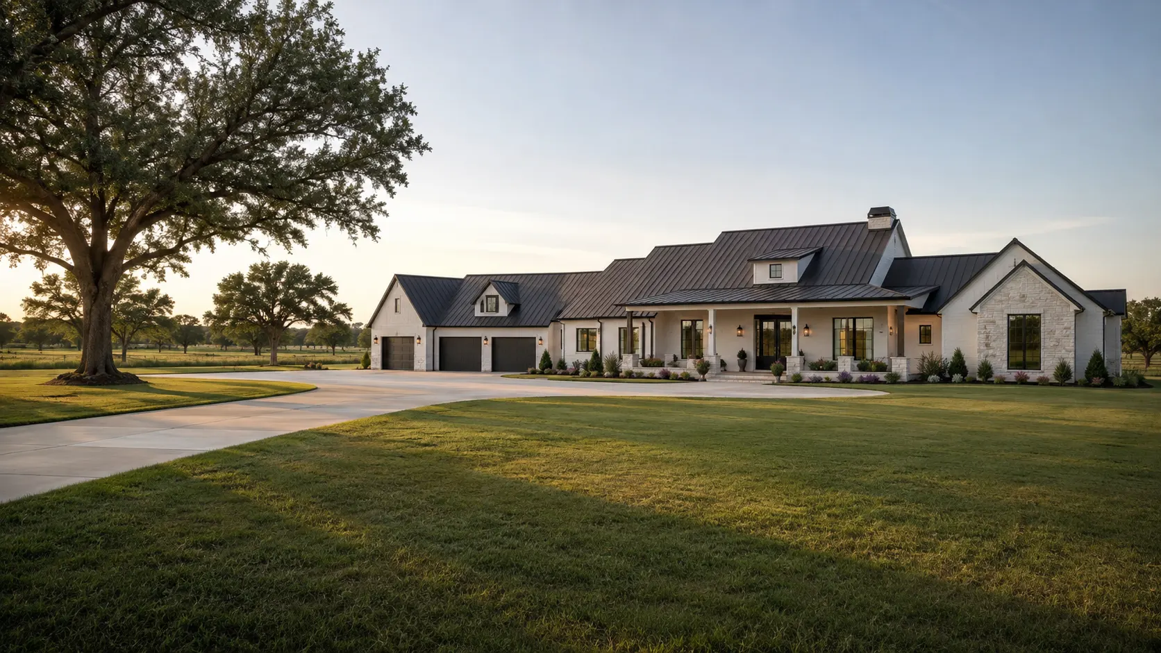 A single-story white brick and limestone custom home with a dark standing-seam metal roof, dormers and a three-car garage, reached by a wide curving drive past a mature live oak on open lawn near Simonton, Texas